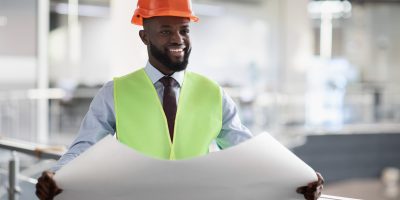 Cheerful black civil engineer in safety workwear holding draft Cheerful young black man civil engineer in safety workwear and orange helmet holding draft, looking at copy space and smiling, architect reading building drawing at construction site