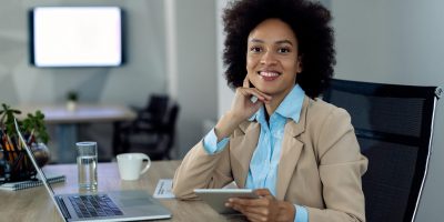 Happy African American businesswoman working on touchpad in the Happy black entrepreneur working on wireless technology at her office desk and looking at camera.